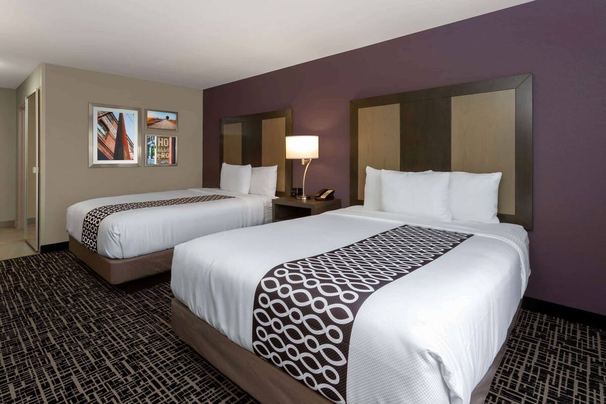 A hotel room with two beds, a nightstand with a lamp, and a door leading to a hallway.