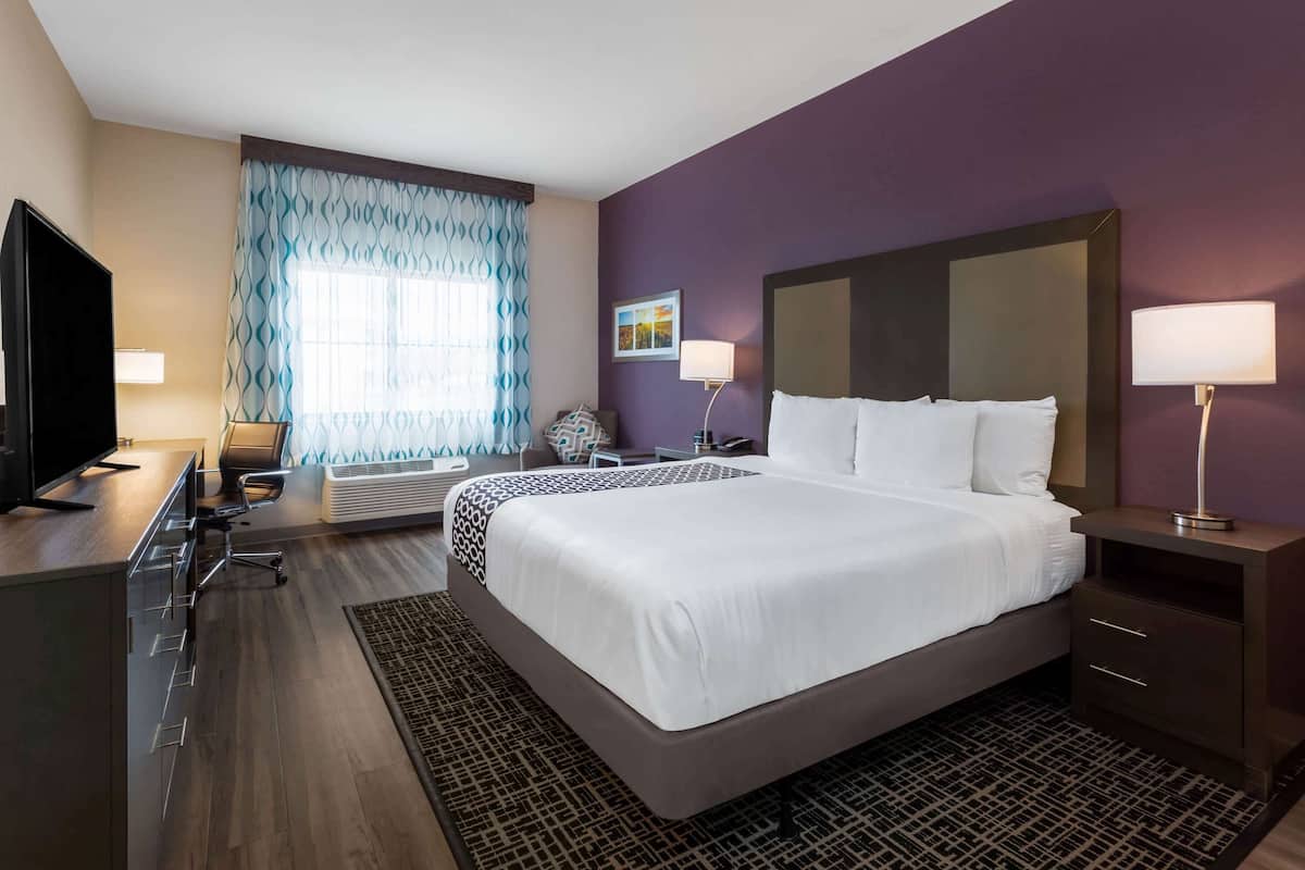 A hotel room with a large bed, a desk with a computer, a flat-screen TV, and a window with patterned curtains.