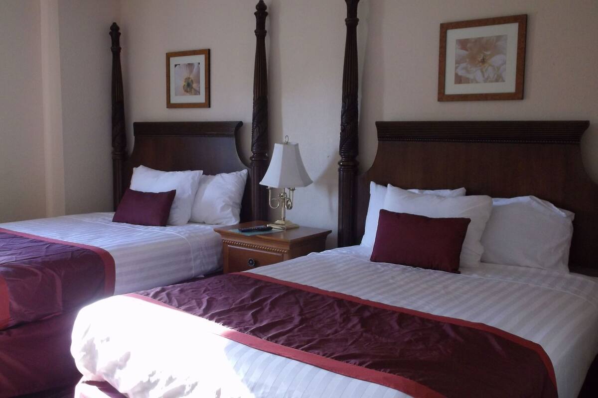 A hotel room with two single beds, each with a wooden headboard, white bedding, and a red accent. There is a nightstand with a lamp between the beds.