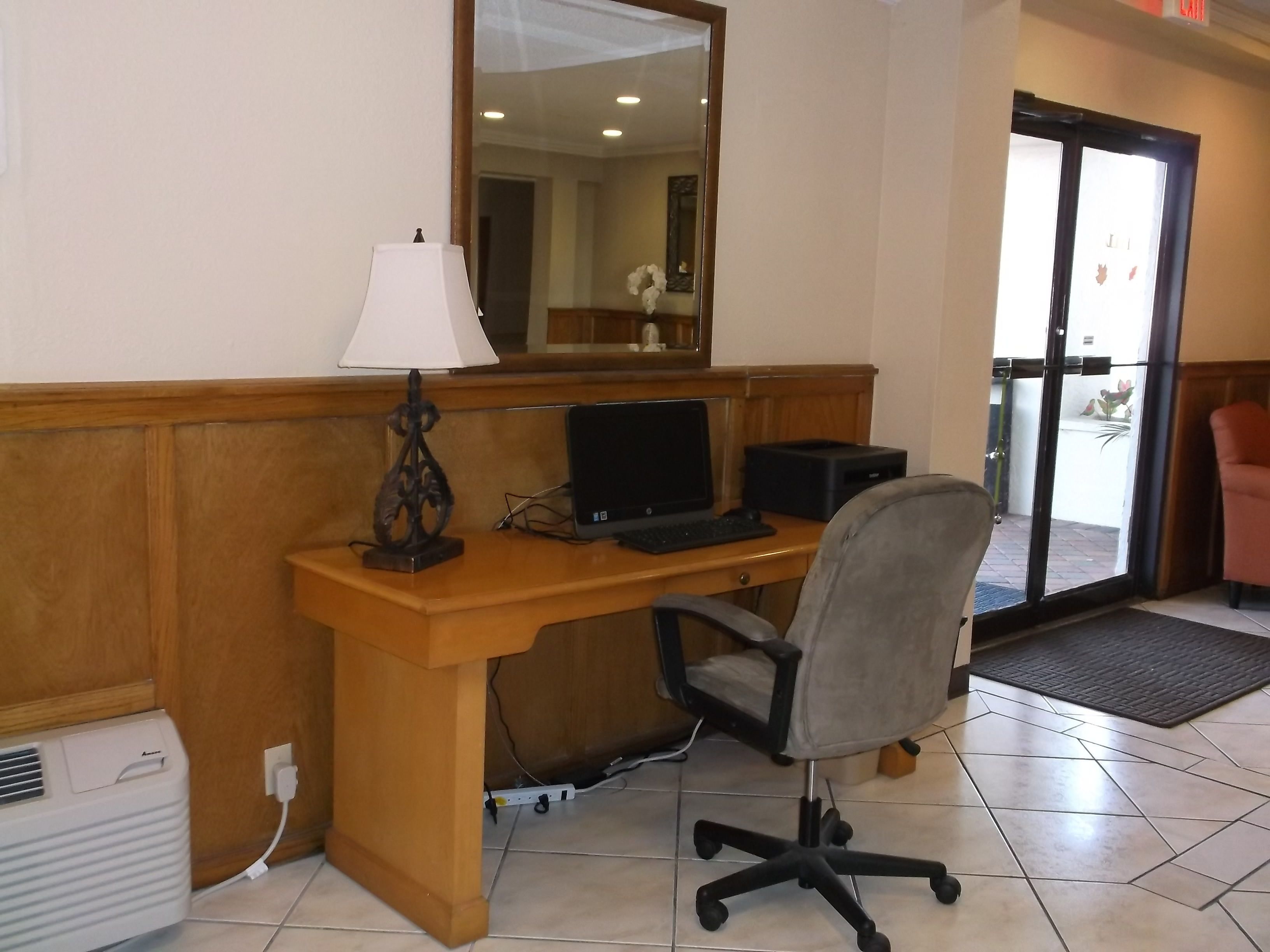 A hotel room with a wooden desk, a computer, a chair, a lamp, a mirror, and a door leading outside.