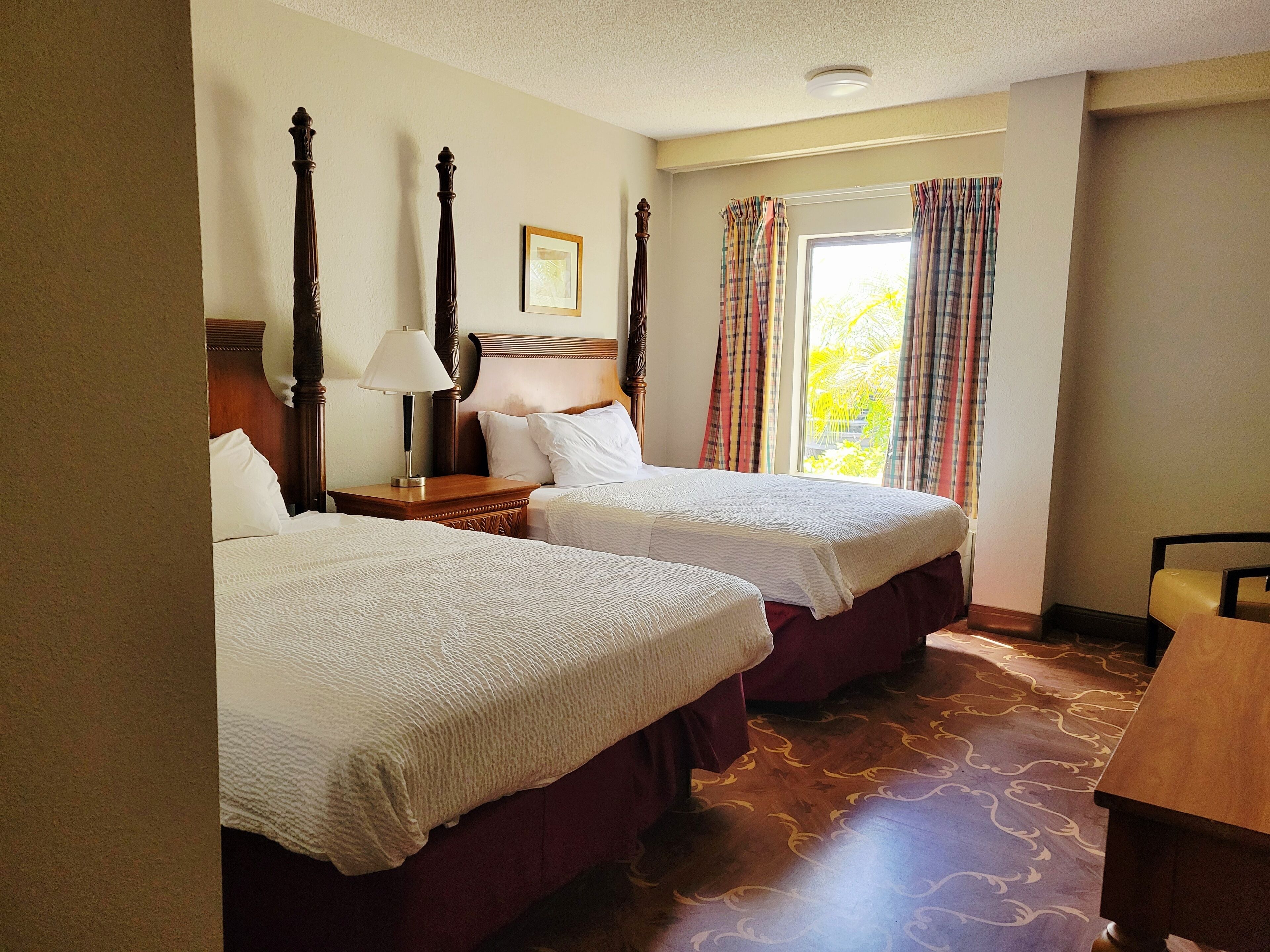 A hotel room with two beds, a wooden headboard, a nightstand with a lamp, and a window with curtains.