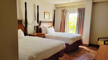 A hotel room with two beds, a wooden headboard, a nightstand with a lamp, and a window with curtains.