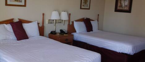 A hotel room with two single beds, each with a wooden headboard, white bedding, and a red accent pillow. There are bedside tables with lamps and a telephone.