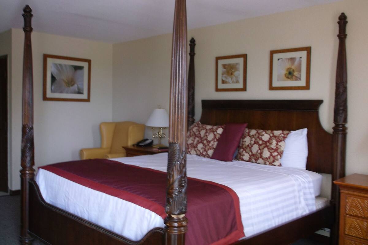 A four-poster bed with a red and white bedspread, a wooden nightstand with a lamp, and a yellow armchair in a hotel room.