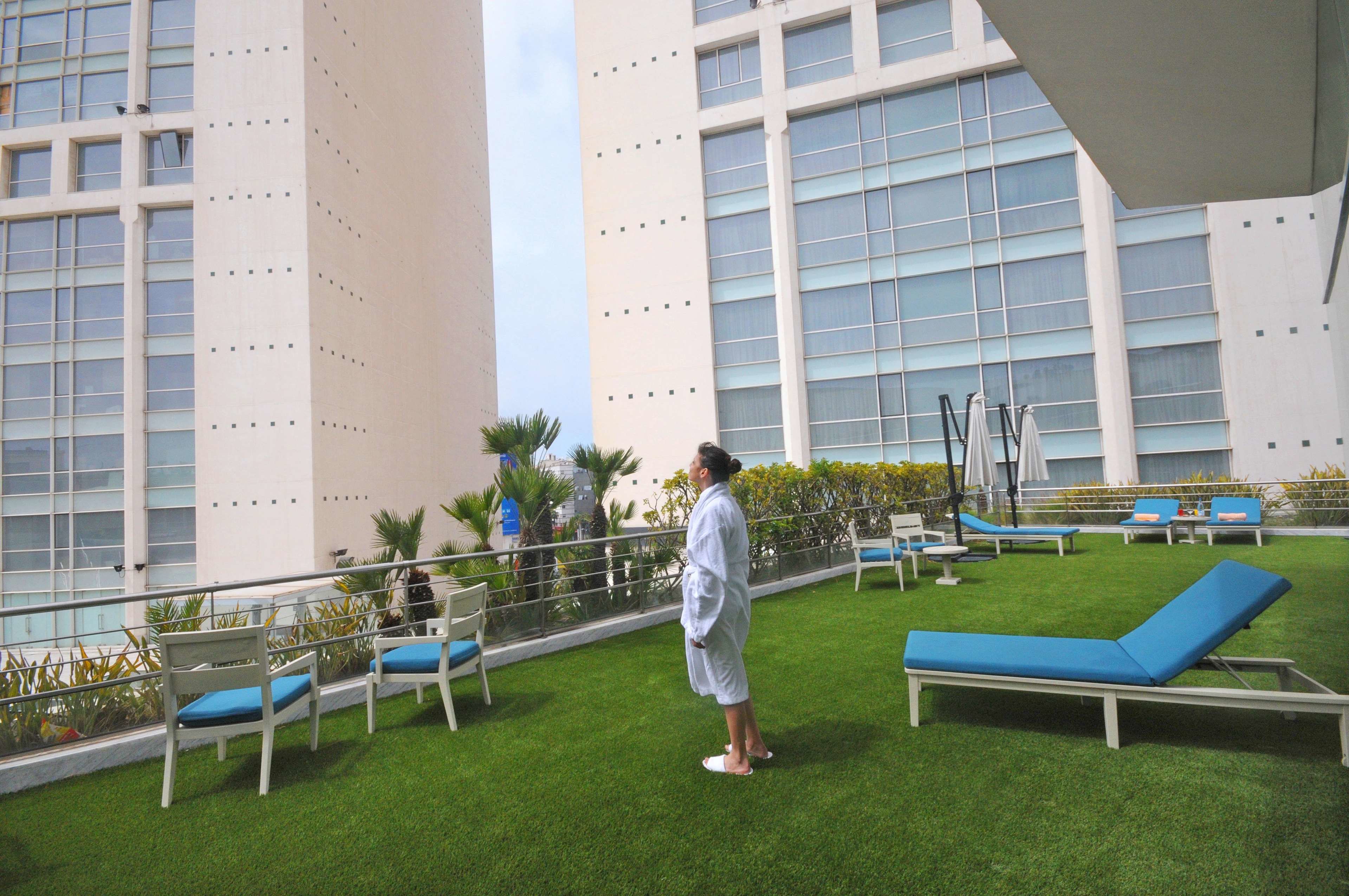 A rooftop lounge with blue lounge chairs and white chairs, surrounded by a low fence and greenery.