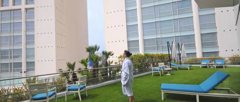 A rooftop lounge with blue lounge chairs and white chairs, surrounded by a low fence and greenery.