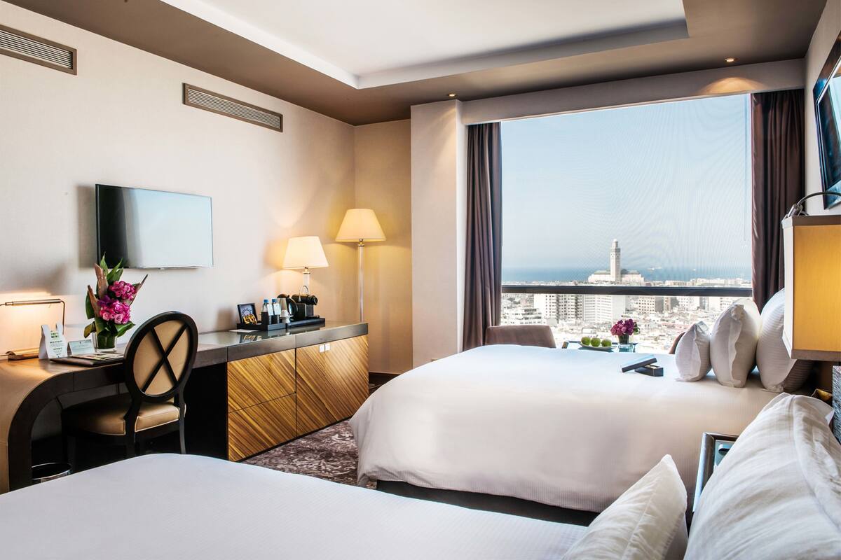 A hotel room with two beds, a desk with a lamp, a TV, and a view of the cityscape.