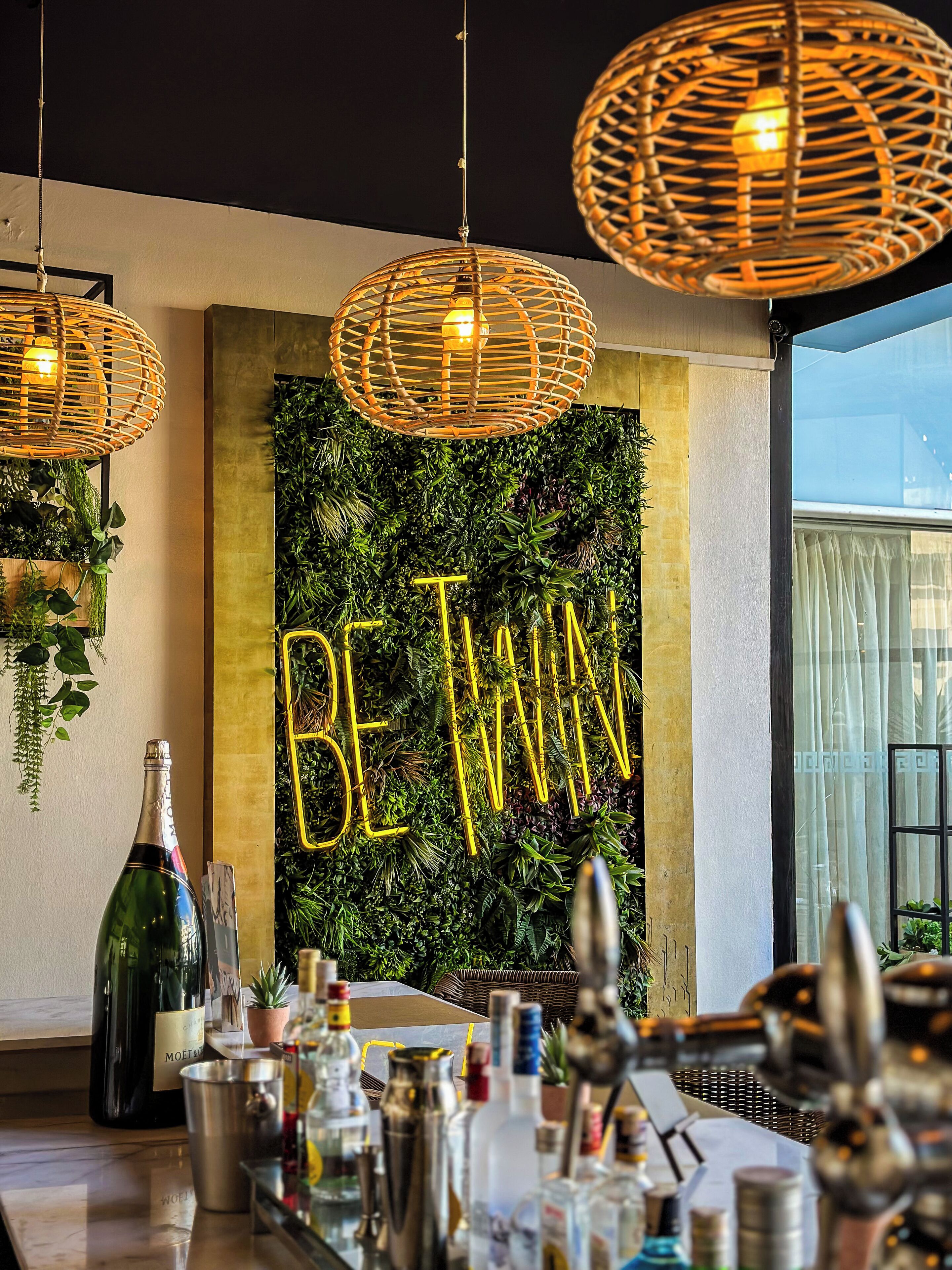 Zona de bar con un letrero 'BE TWINS', lámparas colgantes de mimbre y una variedad de botellas.