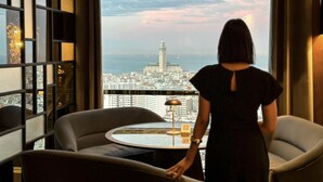 Una mujer con vestido negro de pie junto a una ventana con vista a un paisaje urbano y una torre destacada.