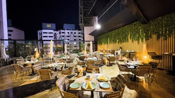 Un espace de restauration en plein air avec des chaises et des tables en osier, un mur décoratif agrémenté de plantes et un bâtiment en arrière-plan.