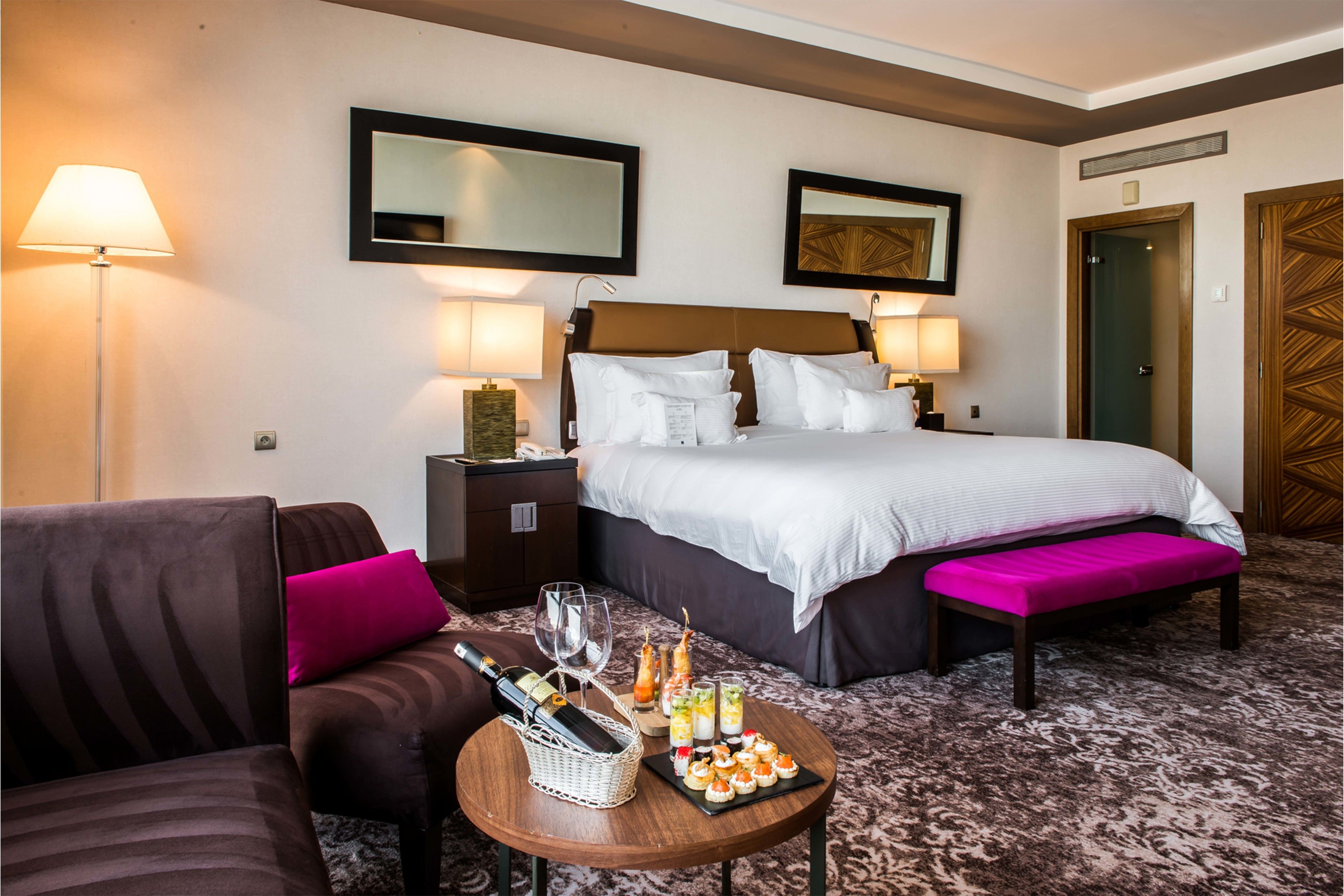 Habitación de hotel con cama, sofá, una mesita con aperitivos y bebidas, y dos lámparas de pared.