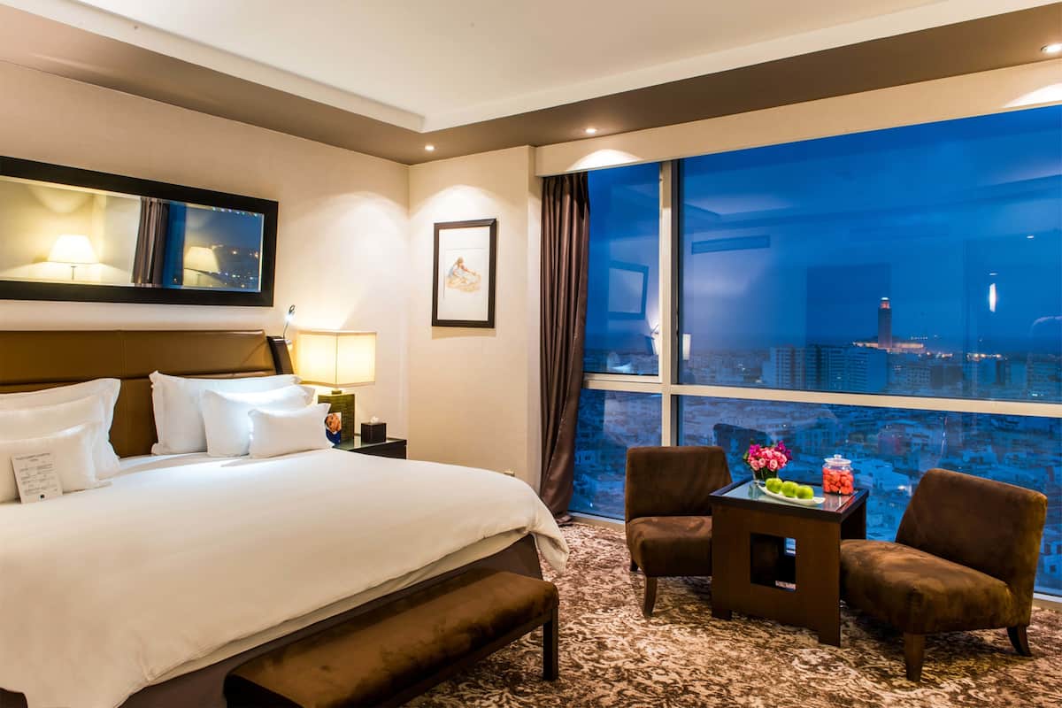 A hotel room with a large bed, a desk with a vase of flowers, two chairs, and a view of a cityscape at night.