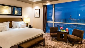 Habitación de hotel con una cama grande, un escritorio con un florero, dos sillas y vista a una ciudad de noche.