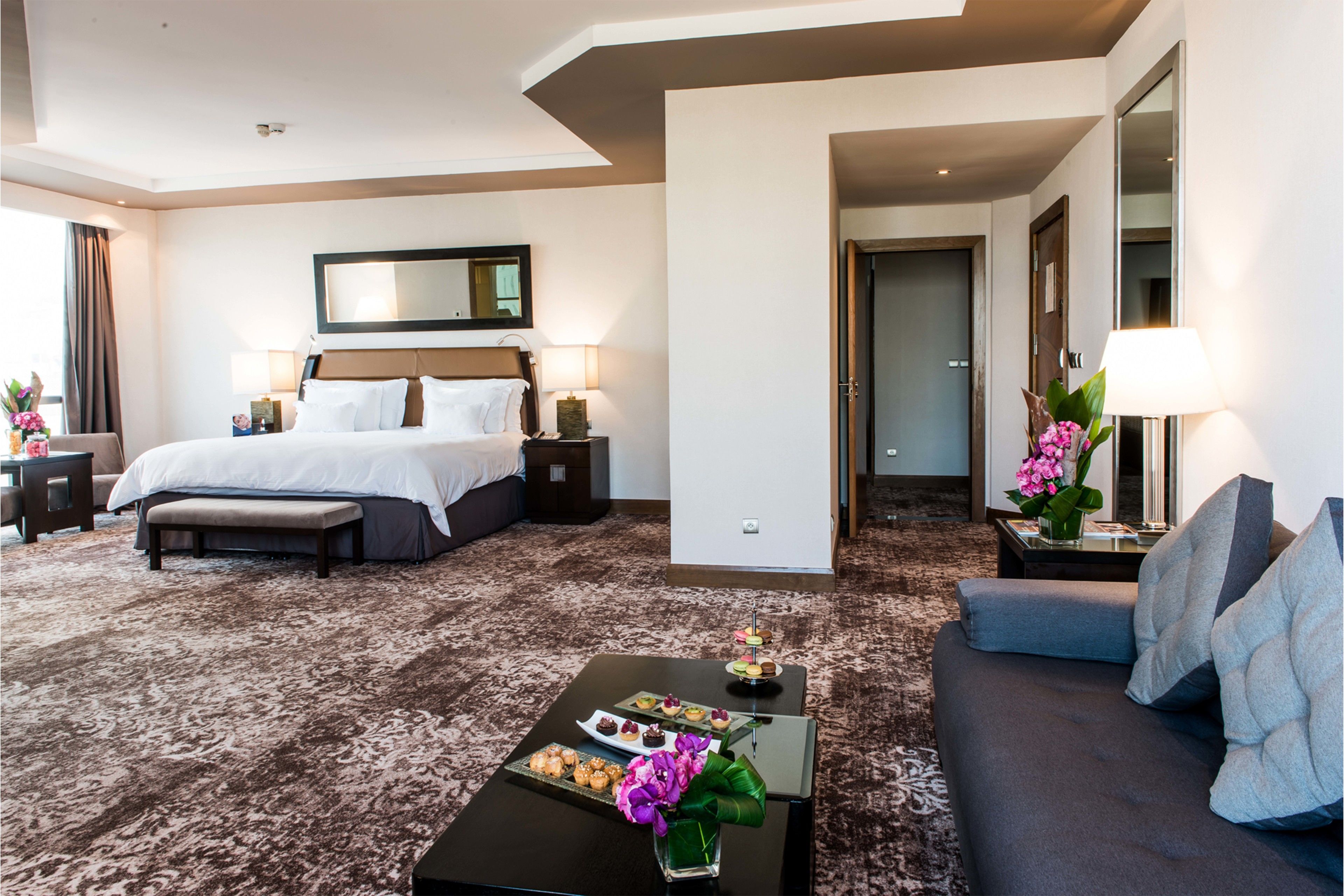 A hotel room with a large bed, a sitting area with a sofa, a coffee table with a tray of pastries, and a mirror above the bed.