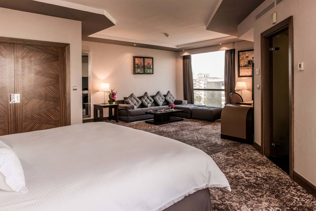 A hotel room with a large bed, a sofa, a coffee table, and a view of the city through the window.