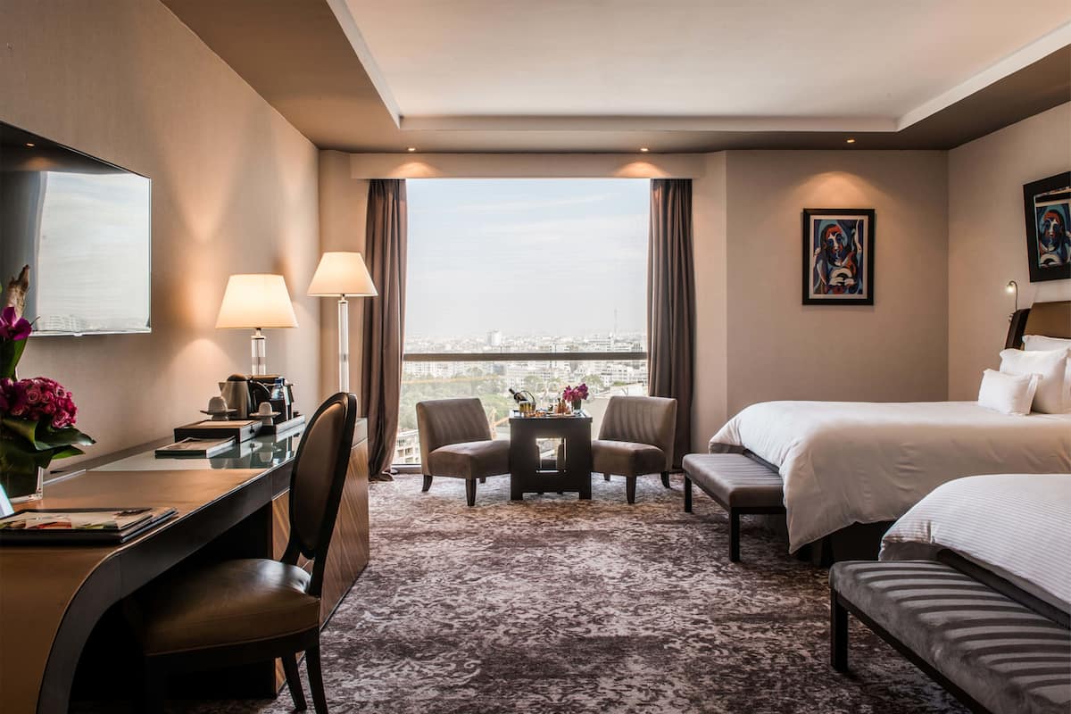 A hotel room with a large bed, a desk with a chair, a small table, and two armchairs. There is a large window with a city view.