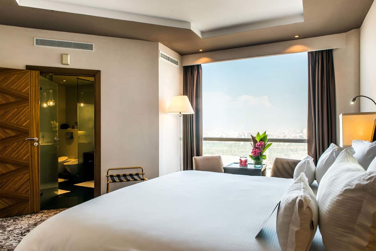 A hotel room with a large bed, a view of the city, a small table with a lamp, and a chair.