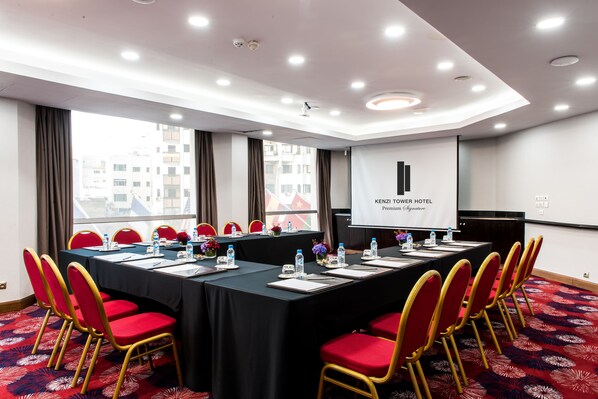 Un salón de conferencias con una larga mesa, sillas rojas y una pantalla de proyector que muestra