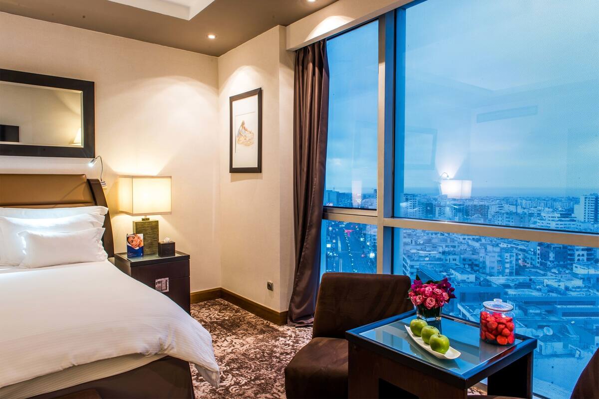 Habitación de hotel con cama, escritorio con un jarrón de flores, mesita de noche con lámpara, ventana grande con vista a la ciudad y un cuadro en la pared.