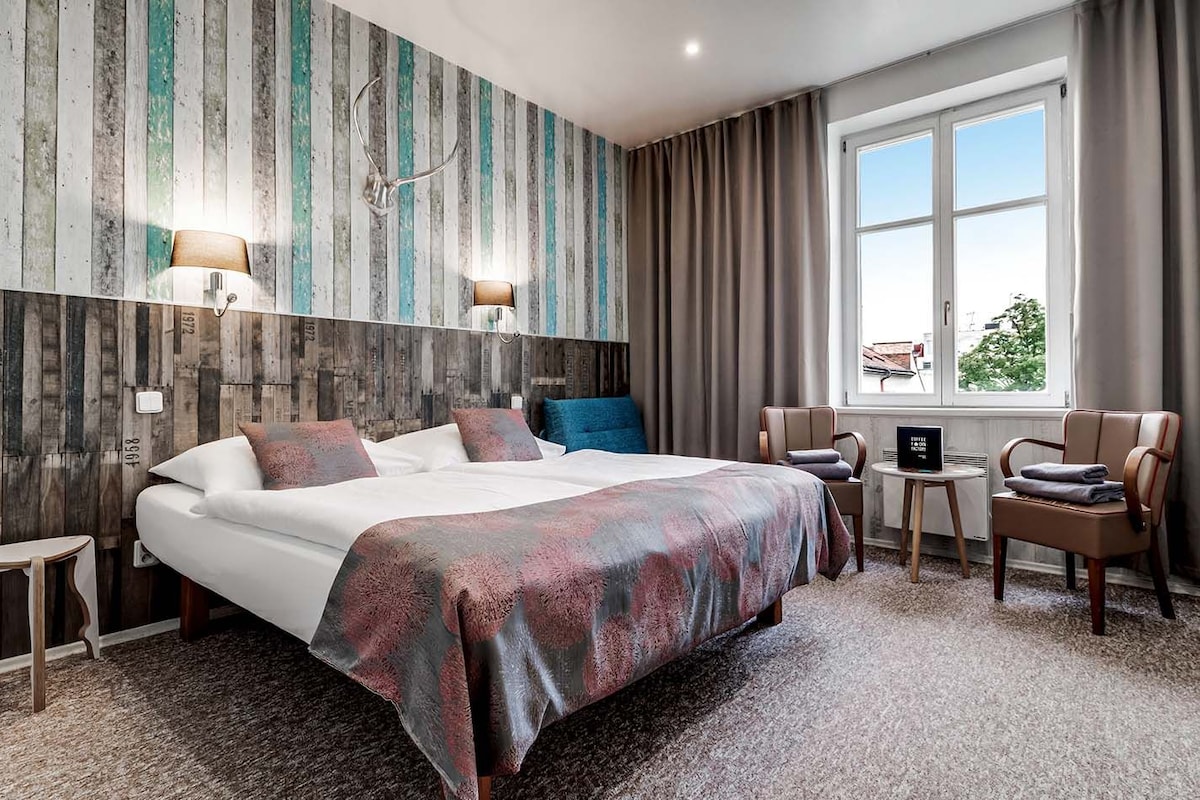 A hotel room with a large bed, a desk, two chairs, a window with curtains, and a wall with vertical stripes.