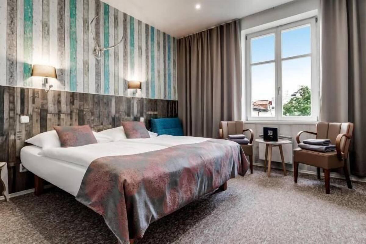 A hotel room with a large bed, two bedside lamps, a window with curtains, a small table, and two chairs.