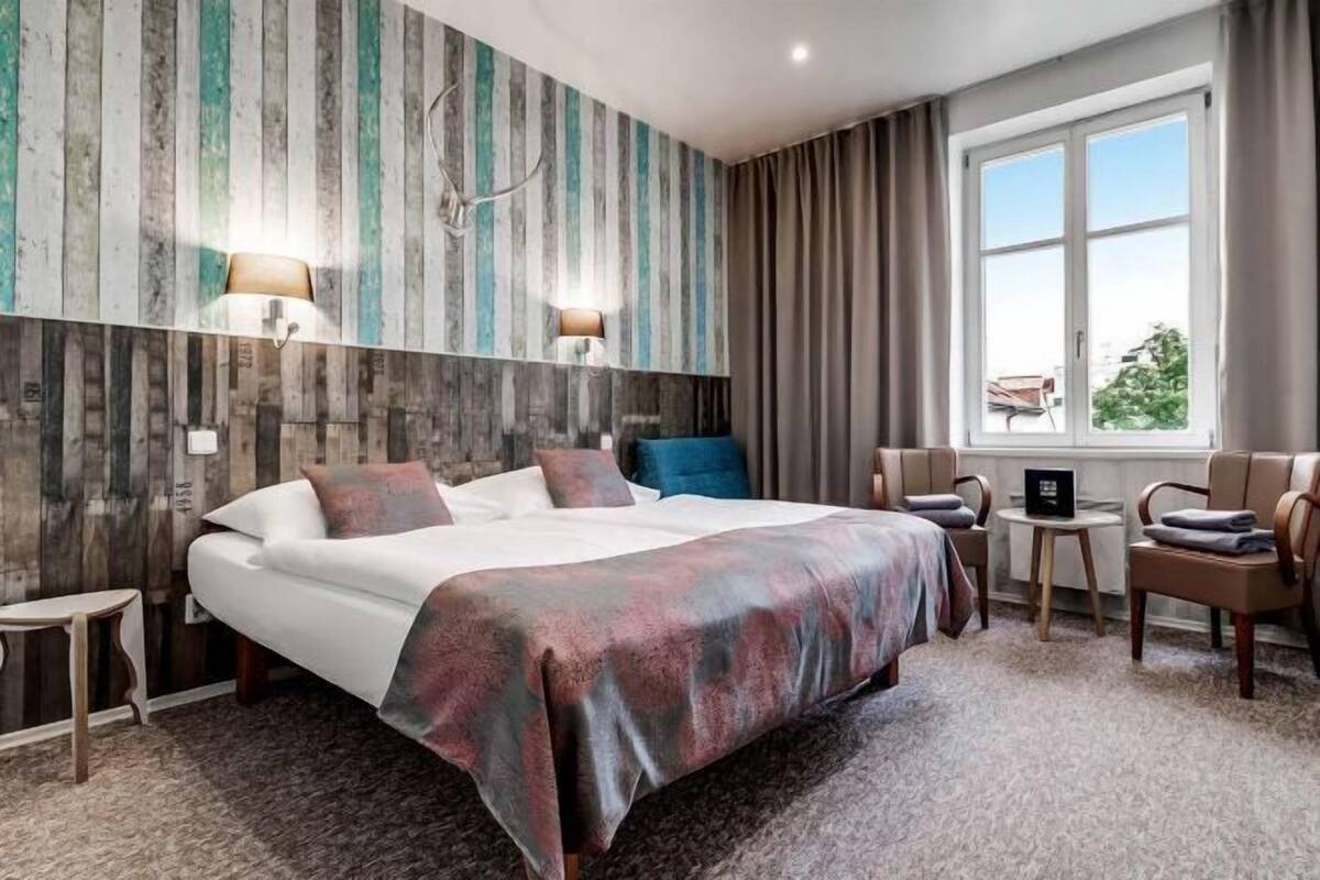 A hotel room with a large bed, two bedside lamps, a small table, a chair, and a window with curtains.
