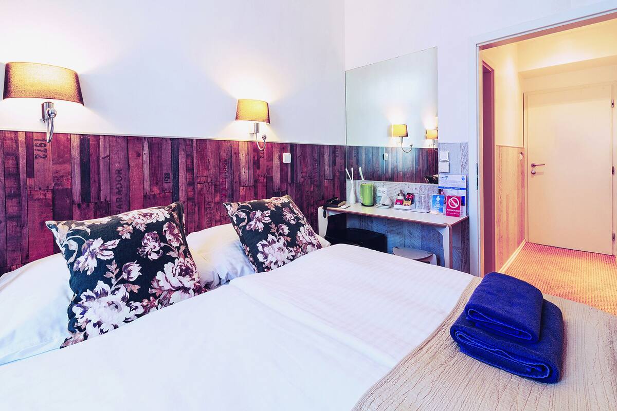 A hotel room with a wooden headboard, a bed with floral pillows, a desk with a mirror, and a blue towel on the bed.