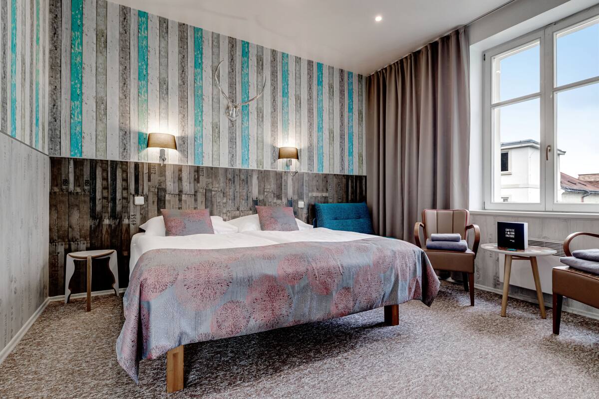 A modern hotel room with a large bed, two wall-mounted lamps, a small table, and a chair.