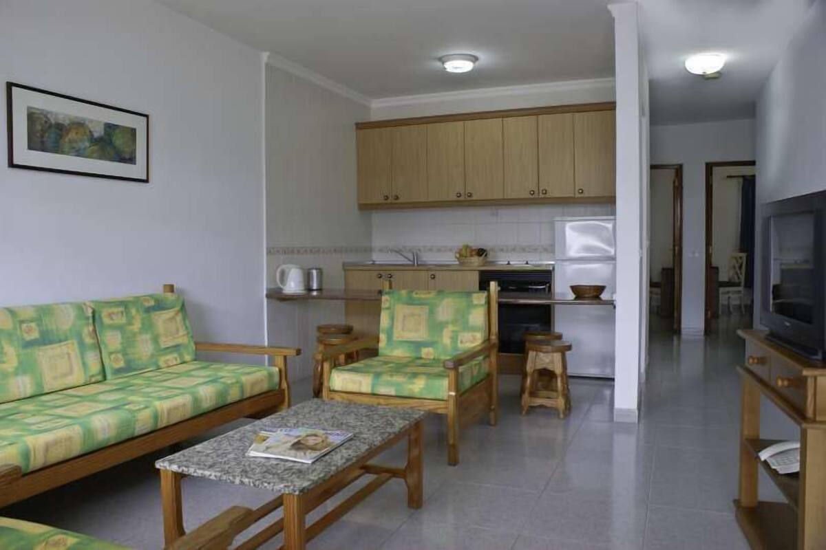A compact living space with a sofa, two armchairs, a coffee table, a kitchen area with cabinets, and a television.