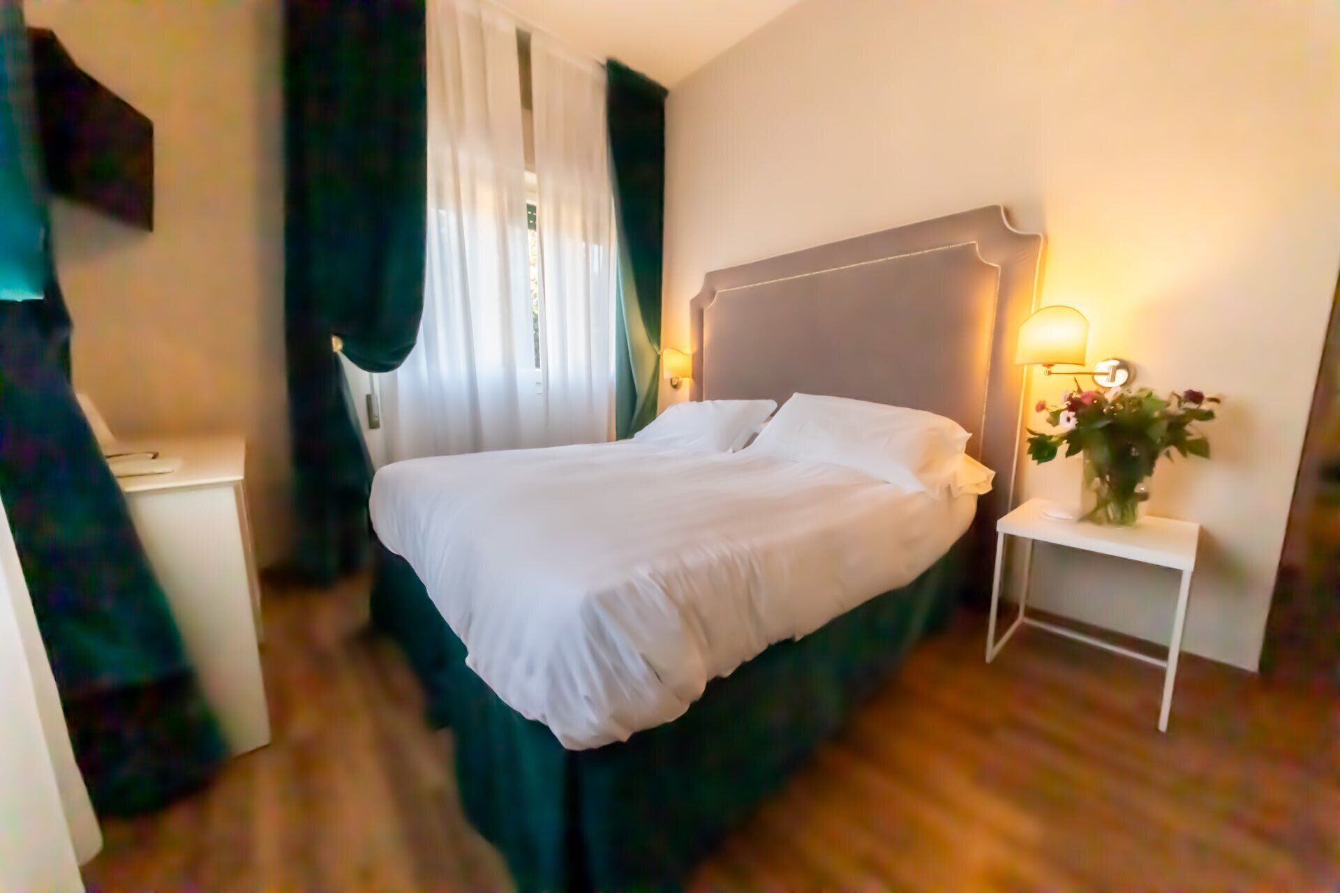 A neatly made bed in a hotel room with a headboard, a bedside table with a lamp, and a small vase with flowers.