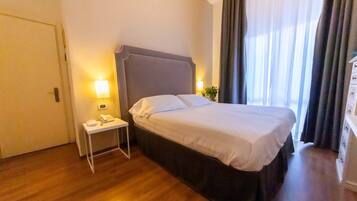 Ein Hotelzimmer mit Bett, Nachttisch, Lampe und einem Fenster mit Vorhängen.