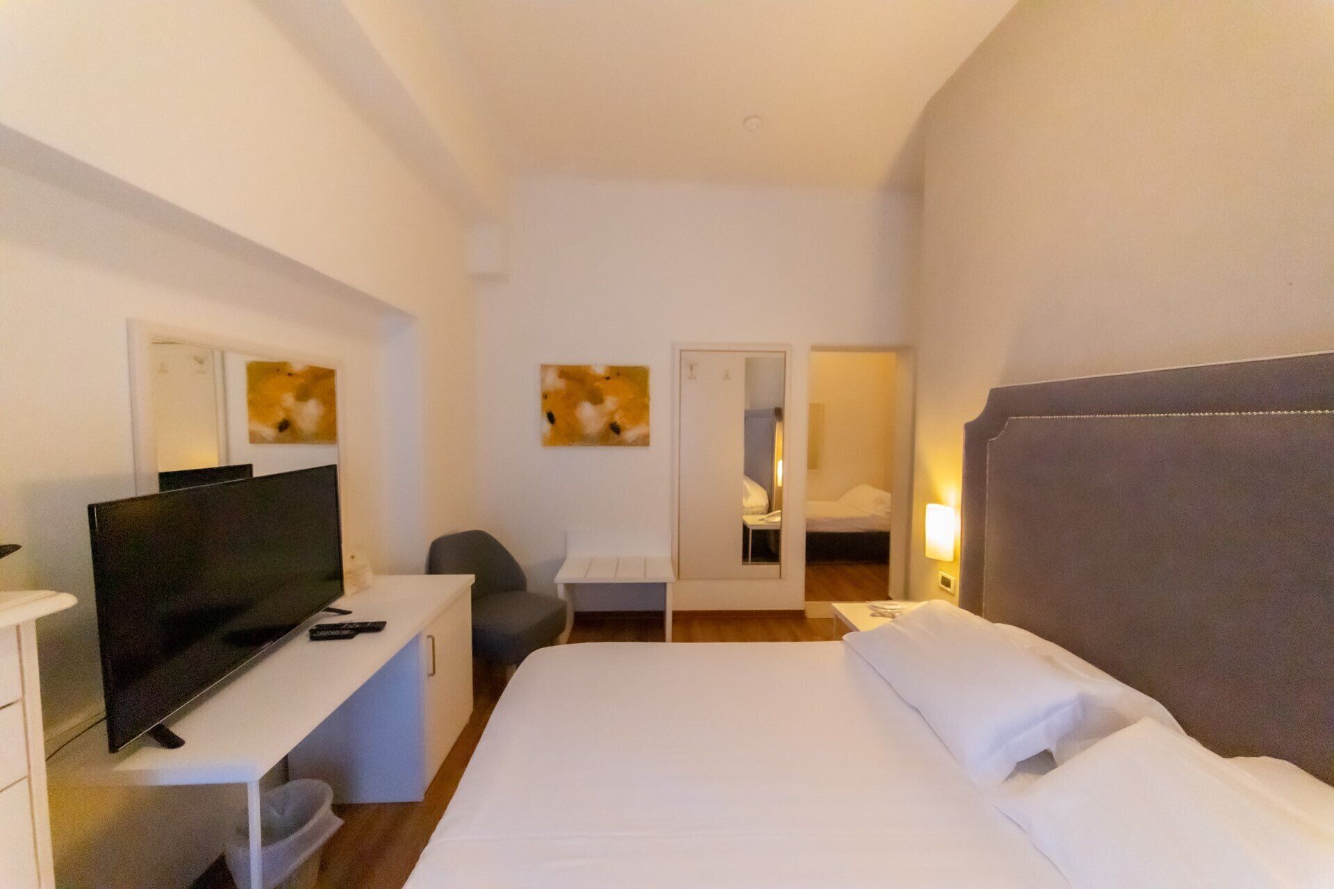 Una habitación de hotel con una cama, un escritorio con televisor, una silla, un espejo y obras de arte en las paredes.