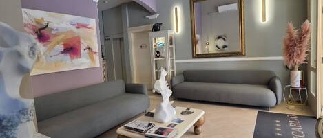 A modern living room with a grey sofa, a coffee table with magazines, a painting on the wall, and a mirror with a gold frame.