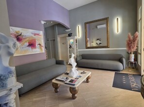 A modern living room with a grey sofa, a coffee table with magazines, a painting on the wall, and a mirror with a gold frame. - Hotel Viscardo (Forte dei Marmi)