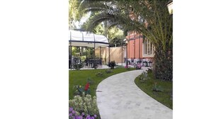 A pathway leading to outdoor seating with tables and chairs, surrounded by greenery and a building in the background. - Hotel Viscardo (Forte dei Marmi)