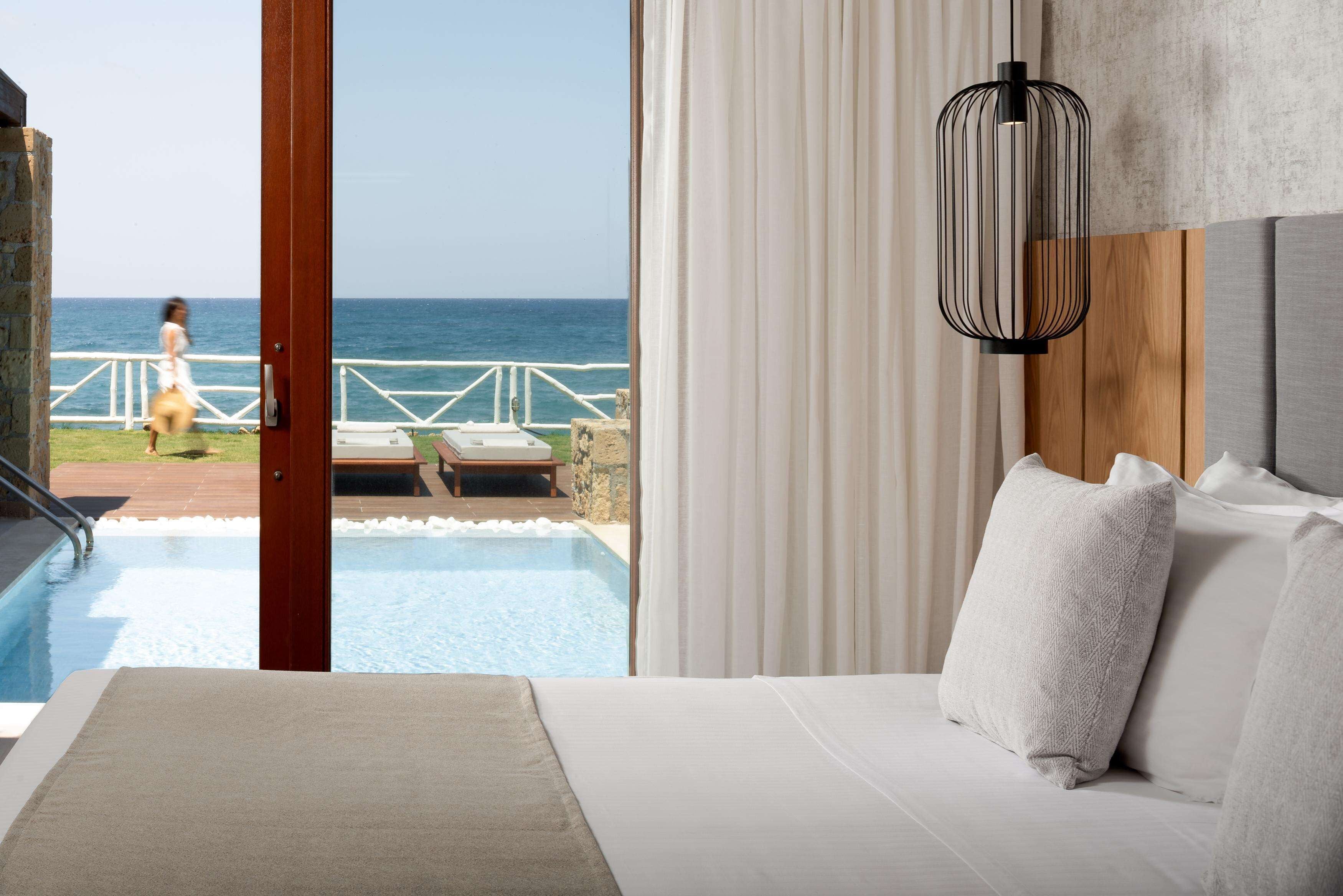 A hotel room with a large bed, a view of the ocean, a pool, and a wooden deck.