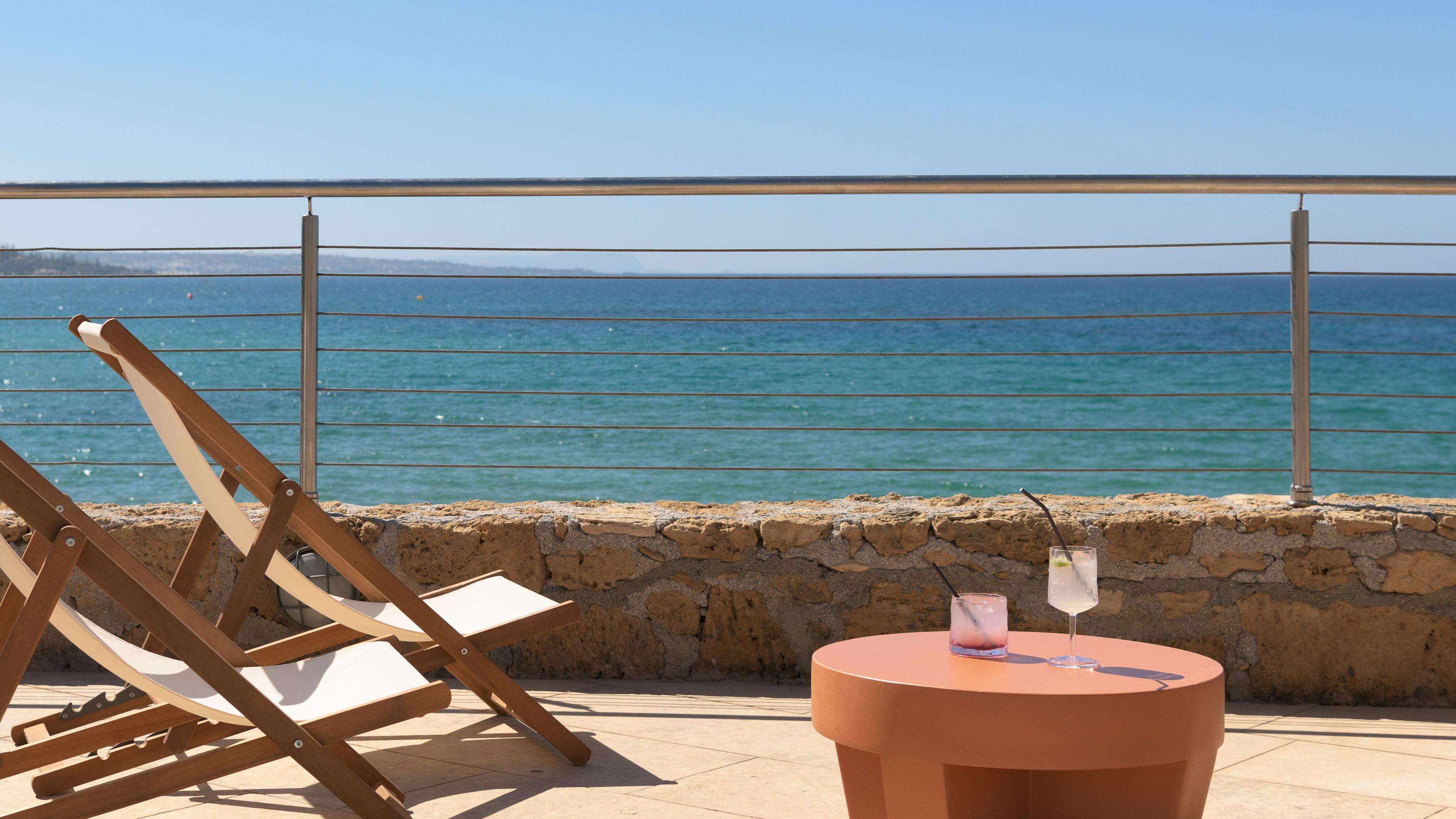 A beachside seating area with a view of the sea, a stone wall, and a round table with drinks.