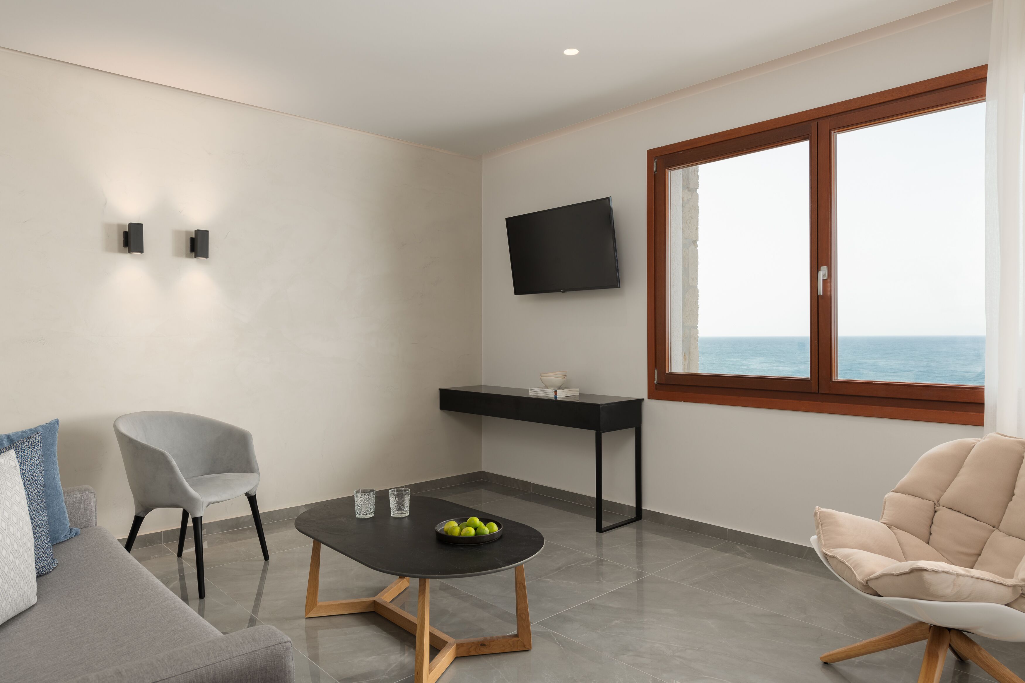 Un salon moderne avec un canapé gris, un fauteuil beige, une table basse noire sur laquelle se trouve un saladier rempli de fruits, une console noire et un téléviseur fixé au mur.