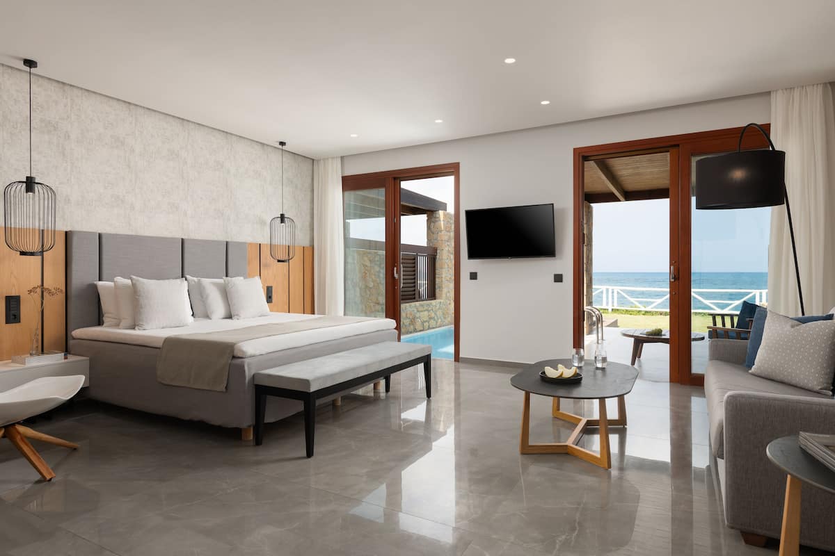 Quarto de hotel moderno com uma cama grande, um sofá, uma mesa pequena e uma televisão. Possui uma varanda com vista para o mar.