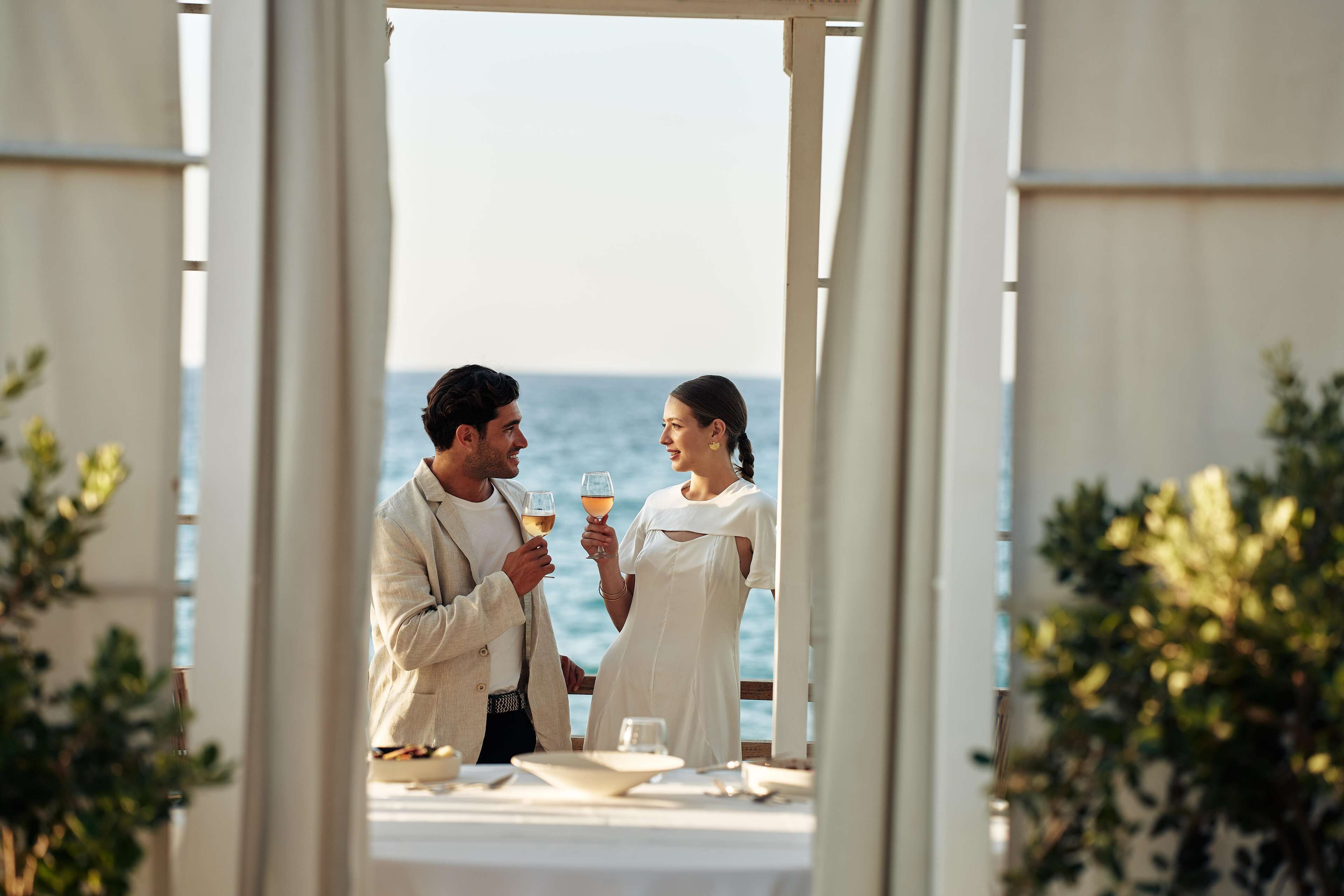 A couple toasting with drinks at a seaside table with a view of the ocean.