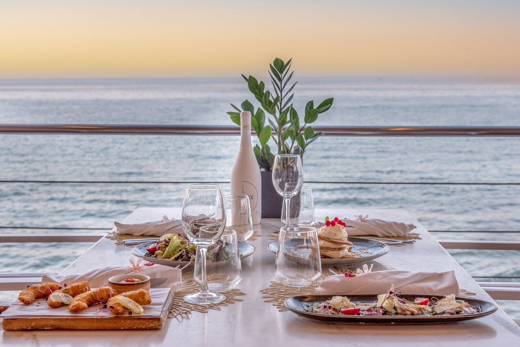 Una mesa lista para una comida con diversos platos, vasos y una botella, con vista a un cuerpo de agua.