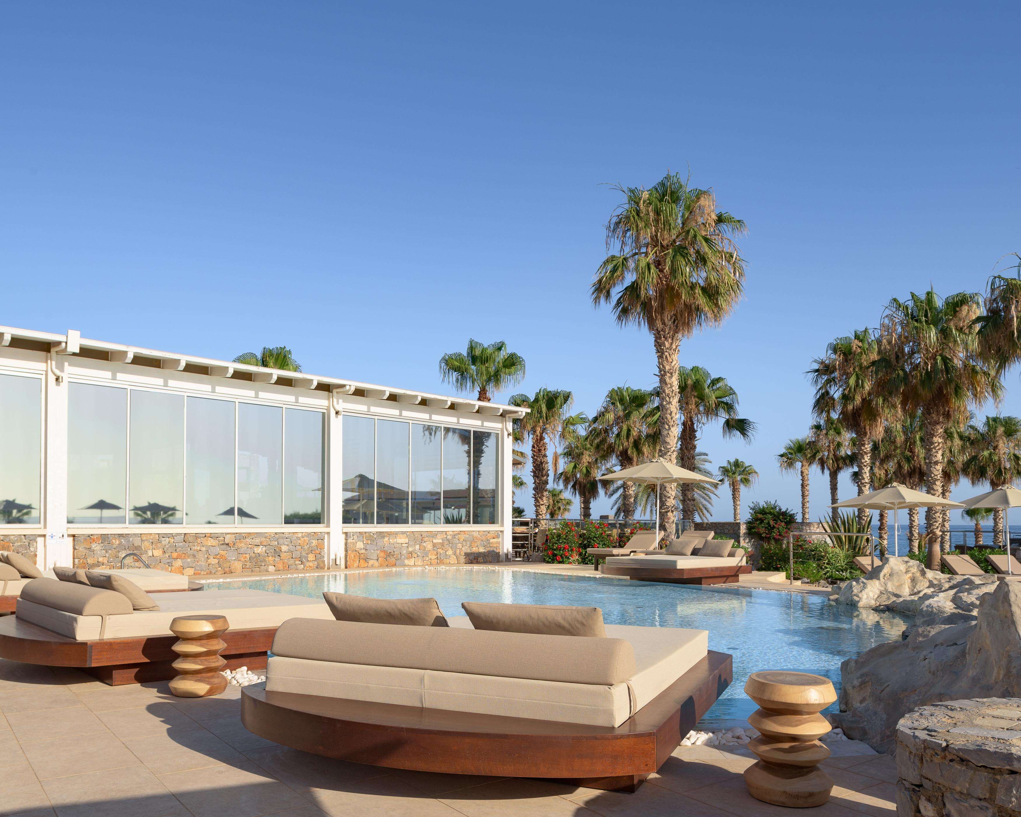 A poolside area with lounge chairs, a glass-enclosed building, and palm trees.