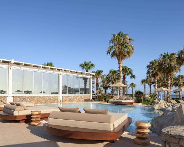 A poolside area with lounge chairs, a glass-enclosed building, and palm trees. - Ikaros Beach Resort & Spa - Adults Only (Hersonissos)