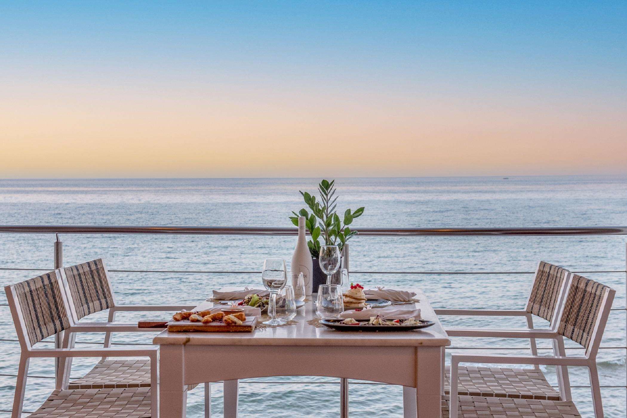 Mesa lista para comer con vista al mar, dos sillas y un florero con follaje.