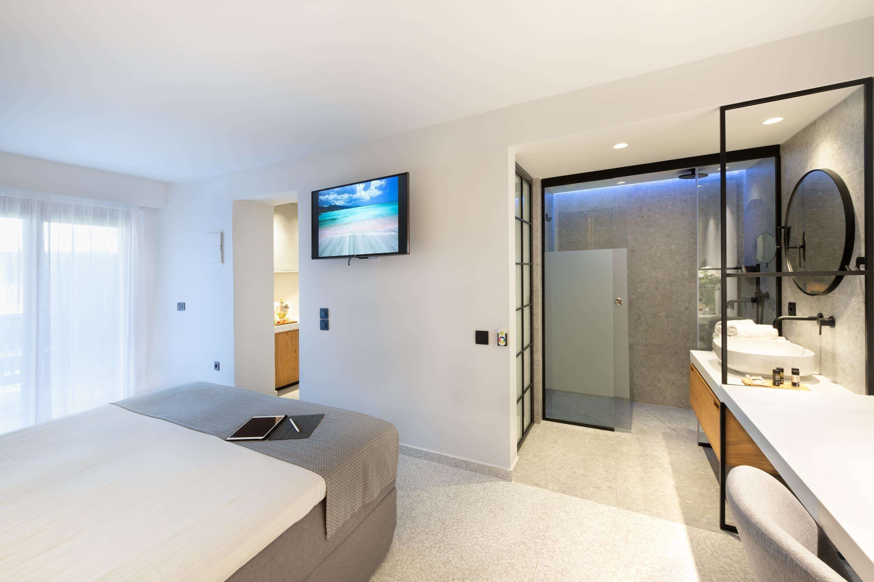 A modern hotel room with a bed, a TV, a bathroom with a mirror and sink, and a view of the outdoors.