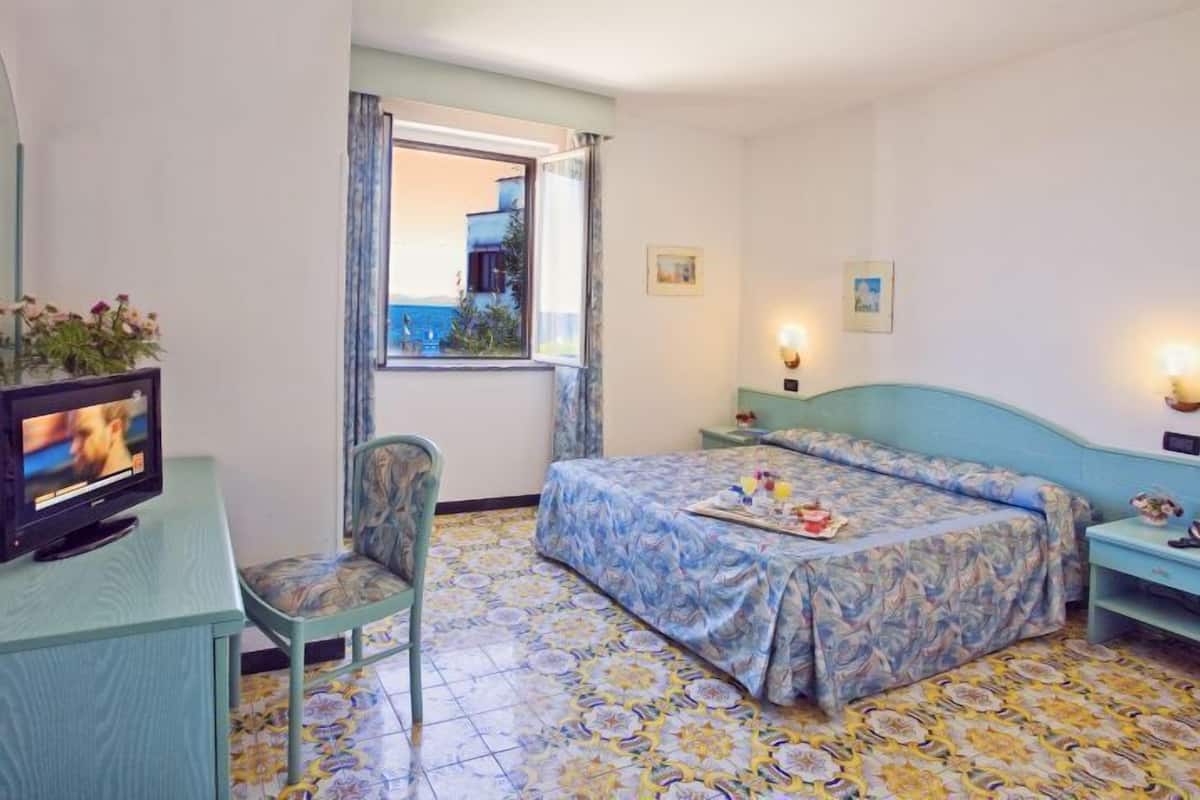 Une chambre d’hôtel avec un lit, une chaise, une télévision, une fenêtre donnant sur un paysage agréable et un sol carrelé.