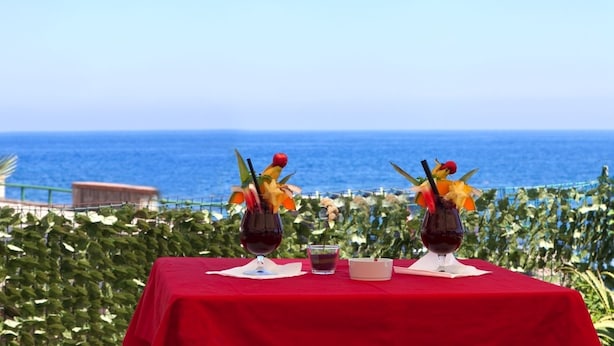 A table with two drinks, a glass of water, and a cigarette pack, set against a backdrop of a clear blue sky and ocean.
