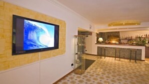 A hotel lobby with a large flat-screen TV mounted on a textured wall, a glass partition, a bar area with stools, and patterned flooring.