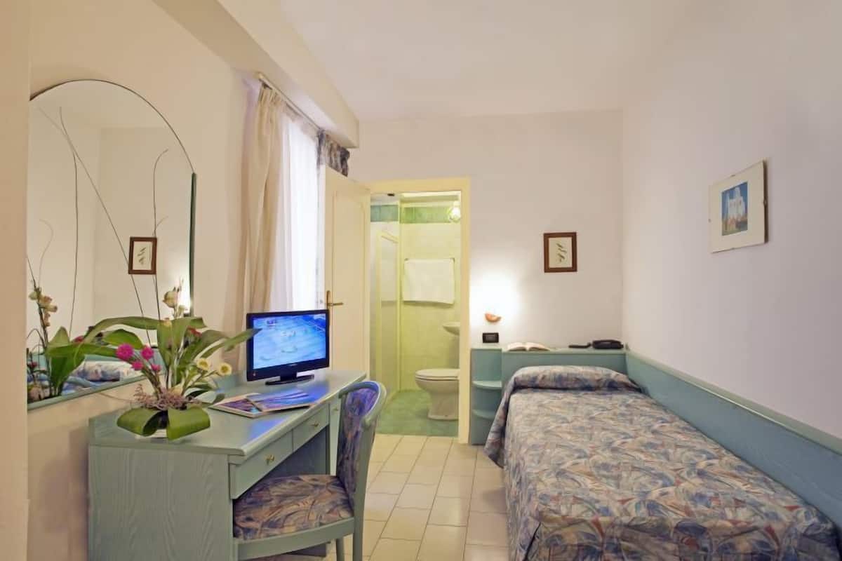 Une petite pièce propre, équipée d’un bureau, d’une chaise, d’un ordinateur, d’un lit et d’une salle de bain visible à travers une porte ouverte.