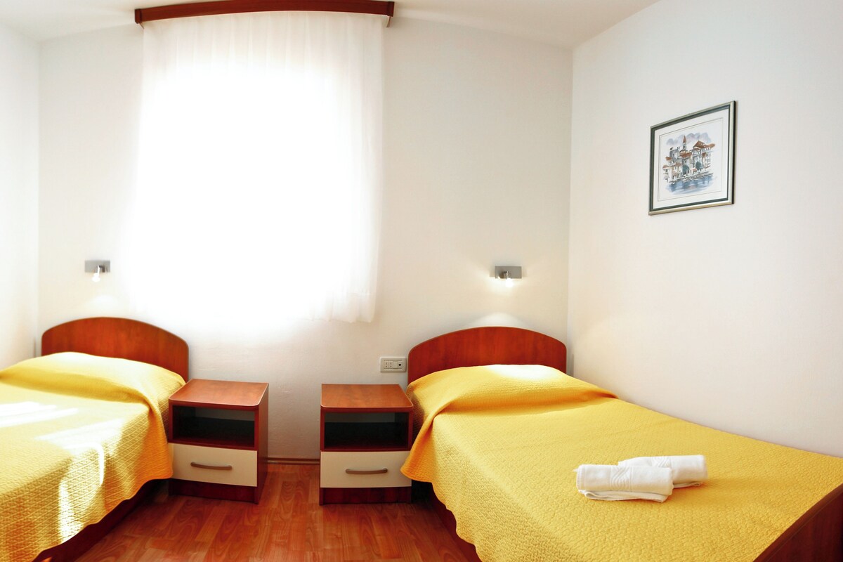 A small, bright room with two single beds, wooden nightstands, and a window with sheer curtains.